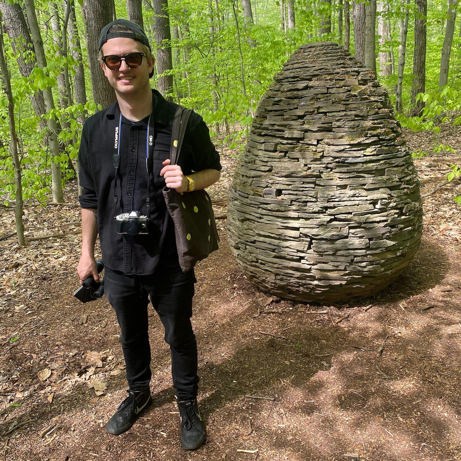 Parker Higgins in sunglasses standing in a forest with a tote bag over his shoulder and a mirrorless camera hanging around his neck. Behind him, a cairn built by Andy Goldsworthy.