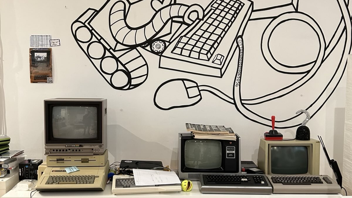 A handful of vintage computers displayed on a table in front of a whimsical mural showing a robot operating a keyboard.